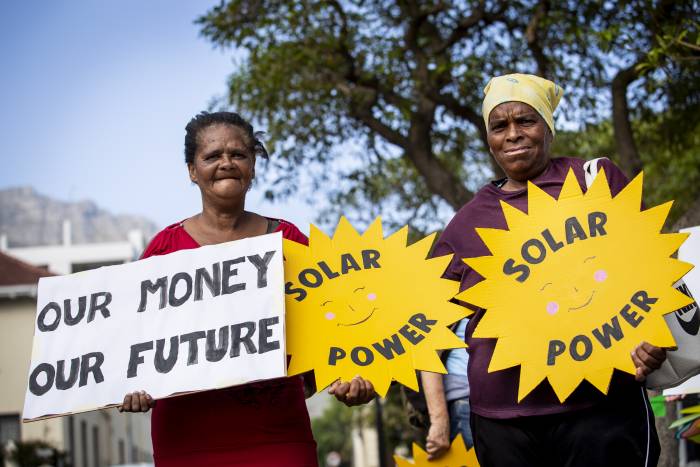 Two women hold home-made signs in front of table mountain that read Our Money, Our Future and Solar Power
